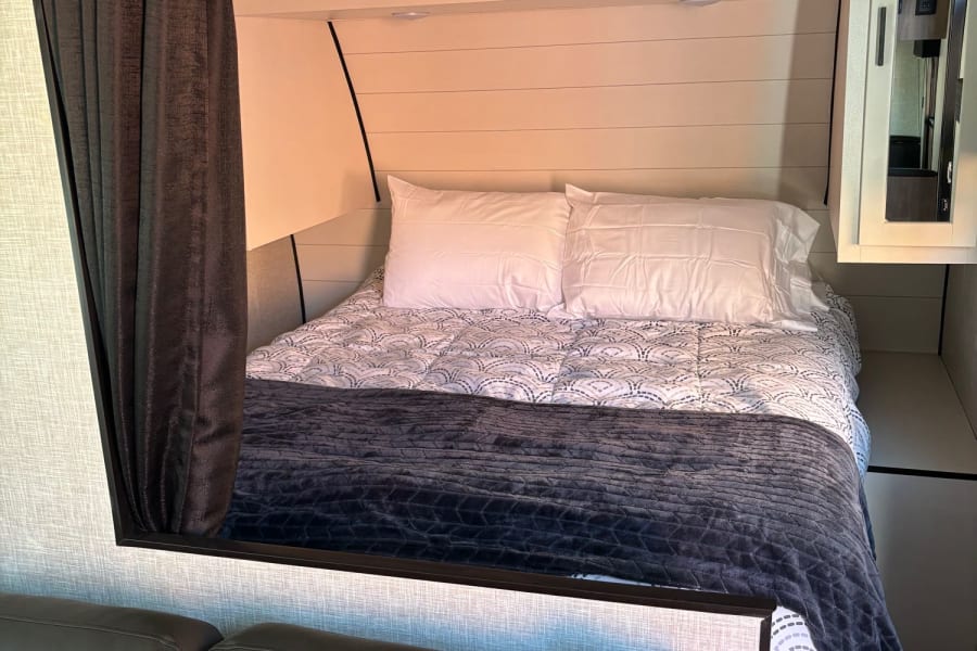 Cozy RV rental bed area with white shiplap walls, dark blanket, and adjacent seating. Ideal for relaxing.