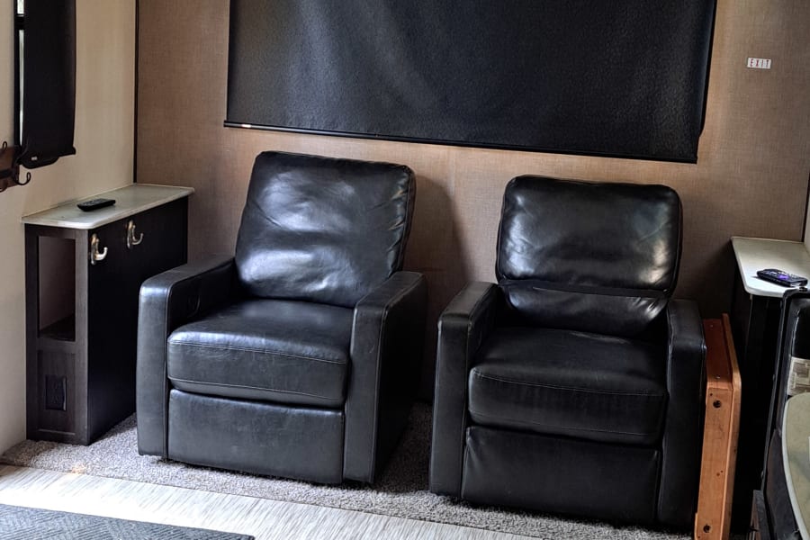 Inside an RV rental: two dark recliners, overhead cabinets, and patterned flooring combining carpet and wood-look vinyl.
