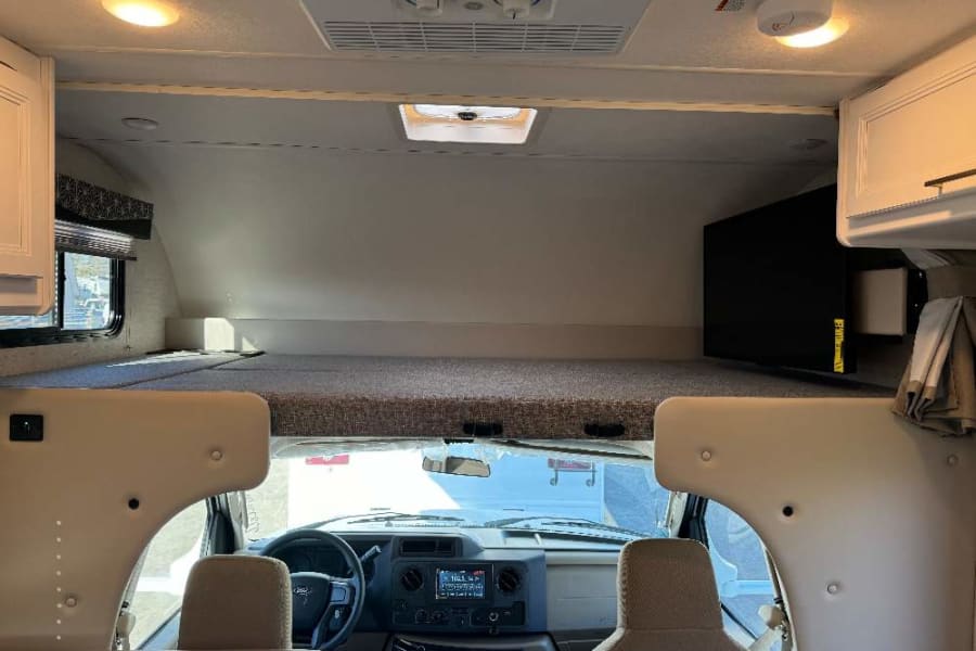 Spacious RV rental interior featuring an overhead bunk bed, wall-mounted TV, and a clear view of the driver's cockpit.