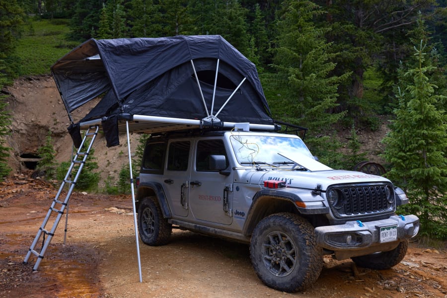 A silver Jeep RV rental with a rooftop tent is ready for camping on a dirt road near an old mine in a forest.