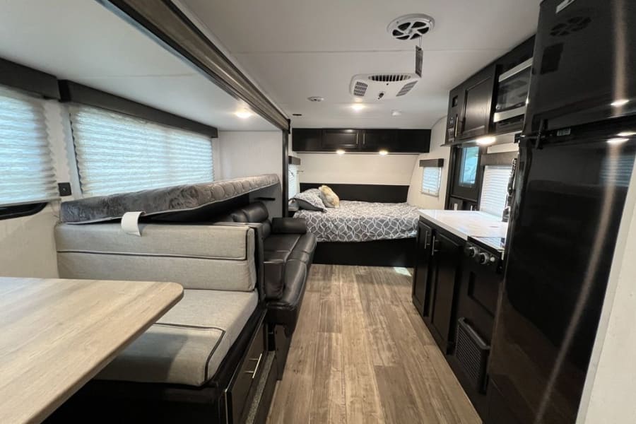 Modern RV rental interior: kitchen with fridge, living area with sofa, dinette with convertible bed, and back bedroom.