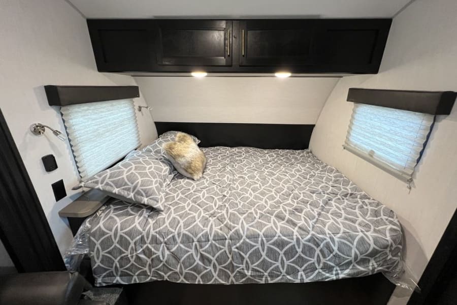 Cozy RV rental bedroom with patterned queen bed, overhead storage, and window blinds for comfort.