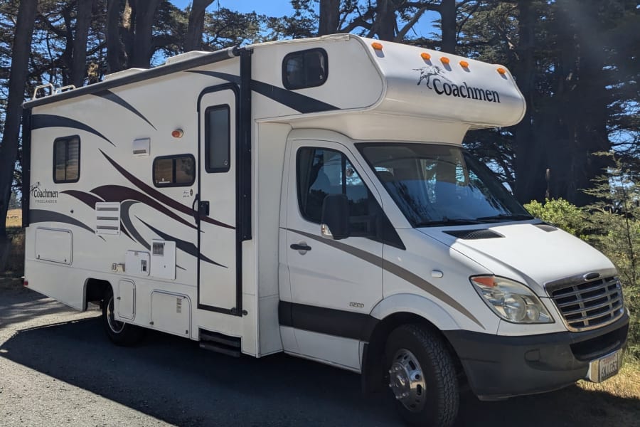 This white Coachmen RV rental is parked among tall trees, ready for an outdoor travel adventure.