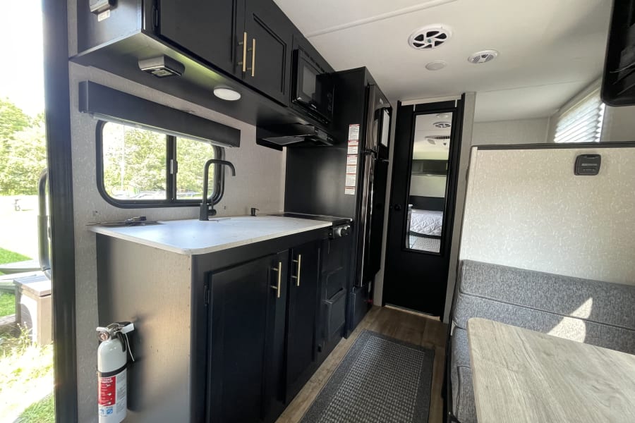 RV rental interior: kitchen with black cabinets, white counters, sink, and an adjacent dining area with gray booth seating.