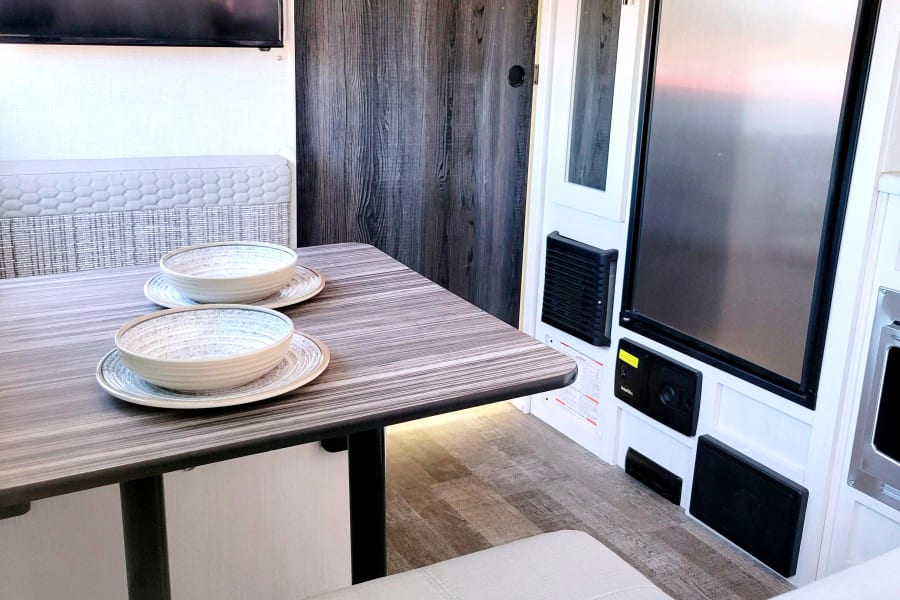 RV rental interior: modern dinette with set plates/bowls, wall TV, fridge, and control panels.
