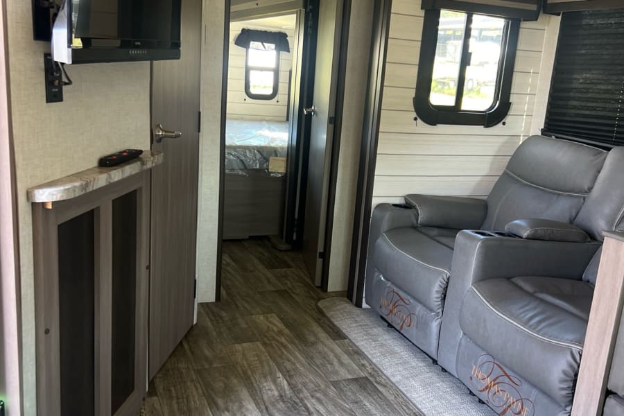 RV rental's inviting living area: gray recliners, TV, and wood flooring, with a glimpse into the connected bedroom.