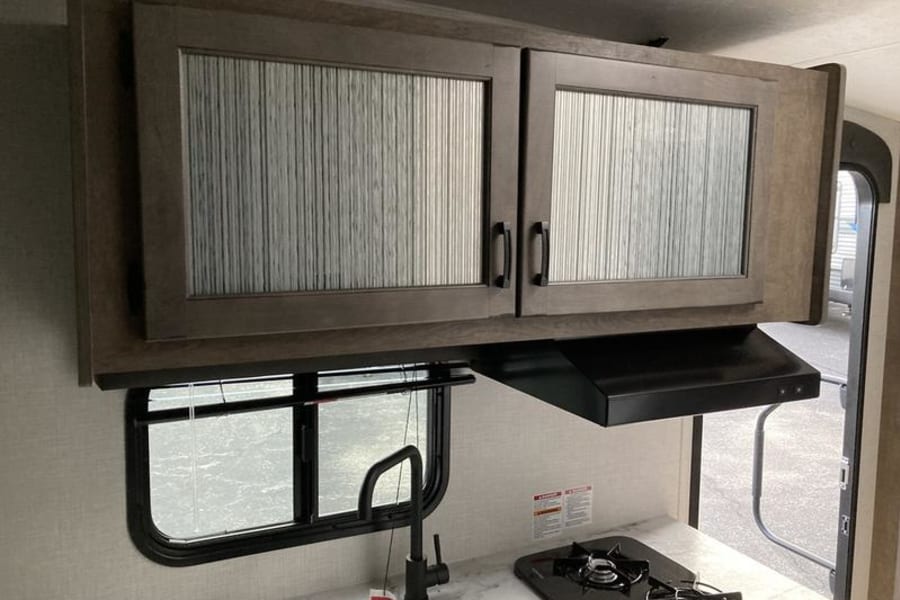 Sleek overhead cabinets with textured panels, a black range hood, stovetop, and window in this RV rental kitchen.