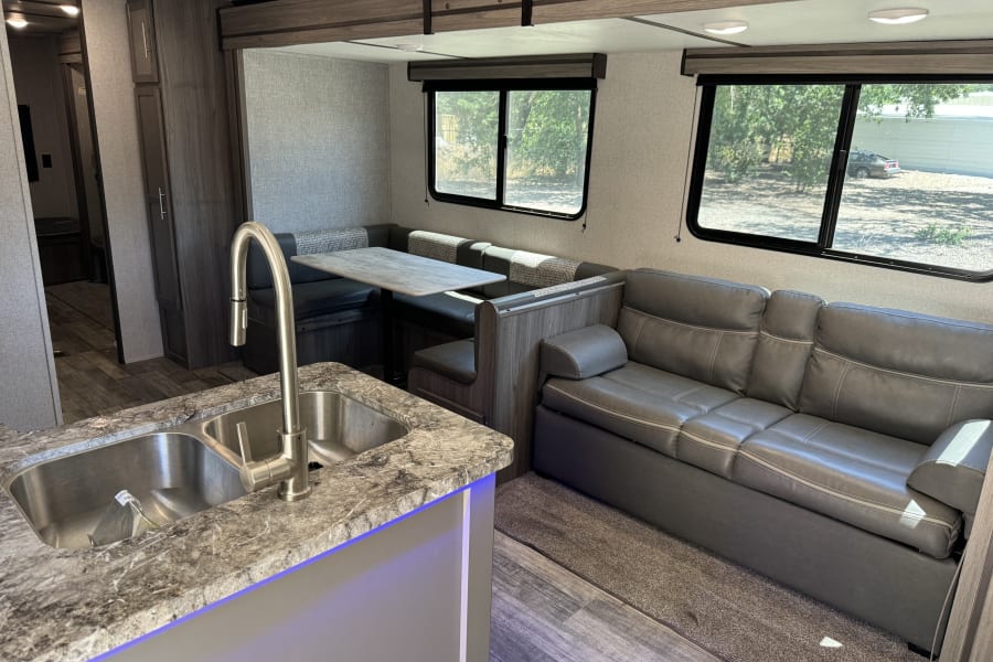 A spacious rv rental interior shows a kitchen sink, dinette with table, and a gray leather sofa. Plenty of light from windows.