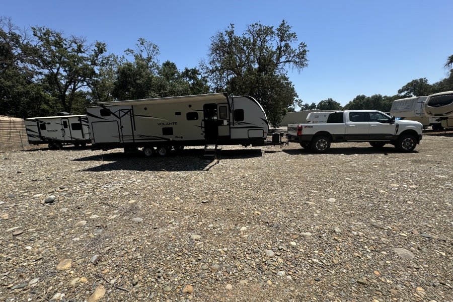 White pickup truck towing a large travel trailer (Volante) in a sunny, gravel RV storage lot, ready for RV rental.