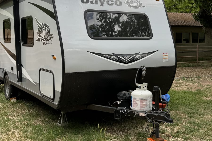 A light gray Jayco travel trailer, ideal for rv rental, parked on grass/gravel with propane tanks on the front.