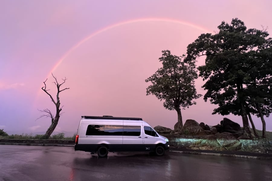 Stunning pink sunset and faint rainbow over an RV rental parked on a wet road, with dark trees and misty mountains.