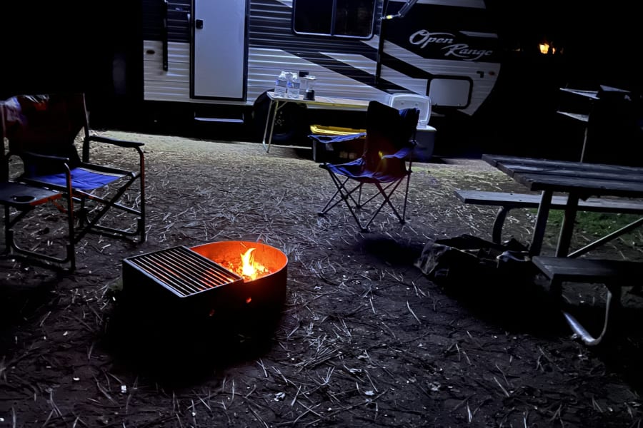 Nighttime rv rental campsite scene with an illuminated travel trailer, glowing campfire, camping chairs, and a picnic table.