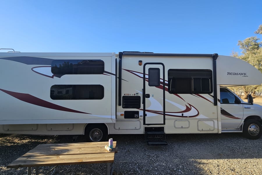 This RV rental is a large motorhome parked at a sunny campsite with its slide-out extended. Two cans sit on a picnic table.