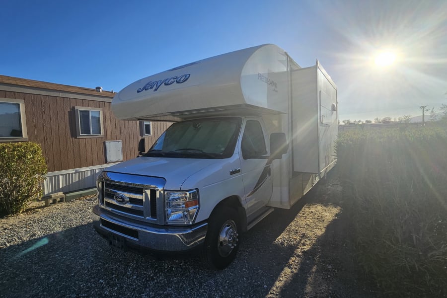 White Jayco Class C RV rental with an extended slide-out, parked on gravel next to a brown mobile home under a bright sun.