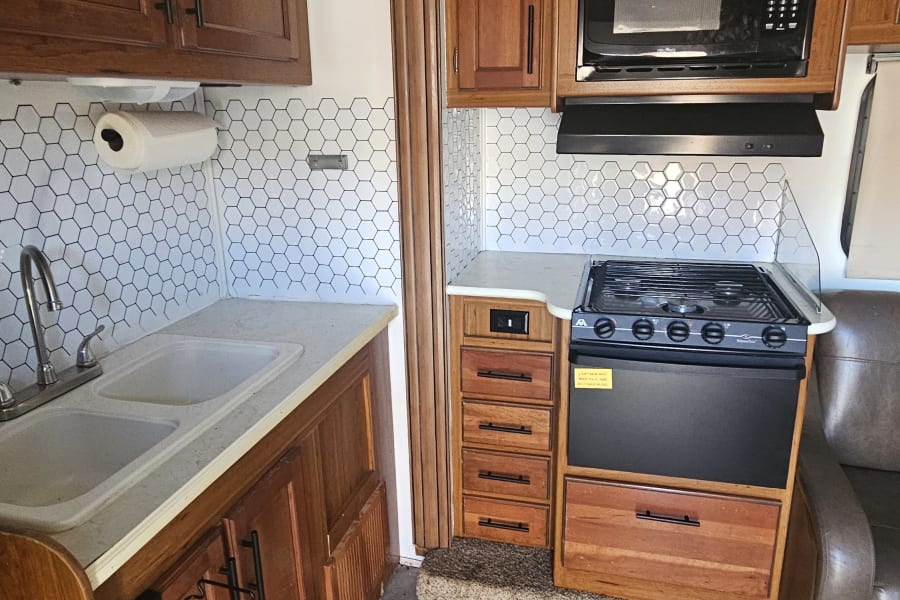 Interior of a clean RV kitchen with wood cabinets, white hexagonal backsplash, double sink, microwave, and stove. Perfect for your rv rental.