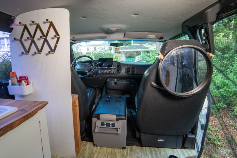 Interior of an RV rental: front cabin with dashboard, portable fridge, and a functional living space with a sink.
