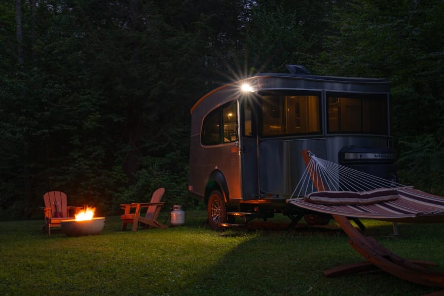 An Airstream rv rental glows at night, with a cozy fire pit, chairs, and hammock in a grassy camping spot.