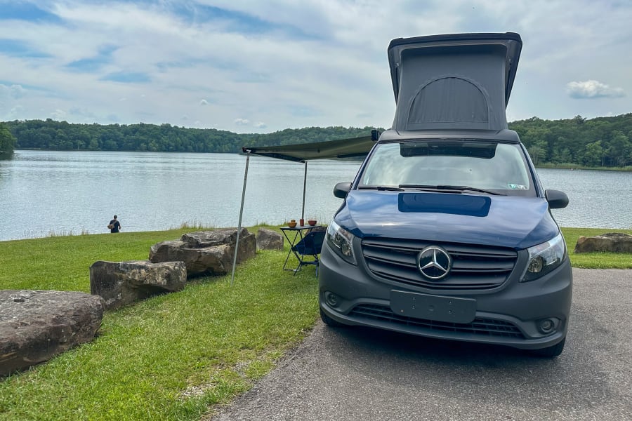 Scenic rv rental: Mercedes pop-top camper van with awning by a tranquil lake, surrounded by green forests, perfect for nature getaways.