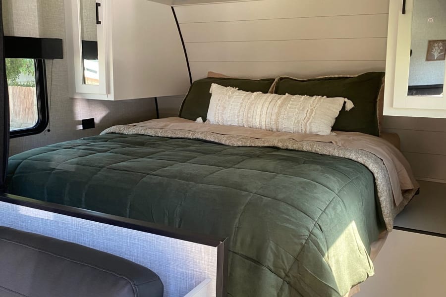 Inviting RV rental bedroom with olive green bedding, overhead storage bins, and mirrored wardrobes. A comfortable travel space.