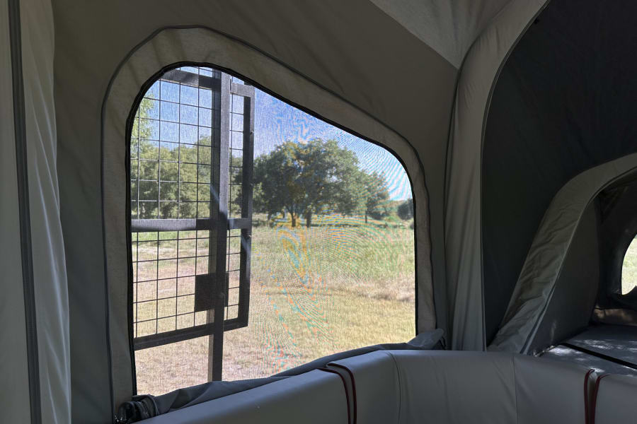 Inside an RV rental, a large screened window with a metal grid frames a view of a grassy field and trees.