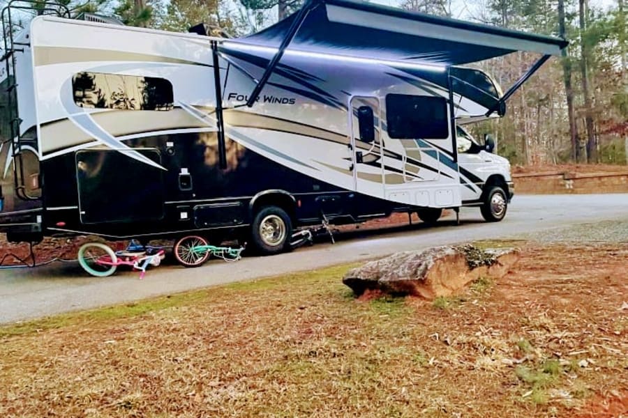 Spacious RV rental with illuminated awning parked on a paved site. Children's bikes are on the ground, perfect for family camping trips.