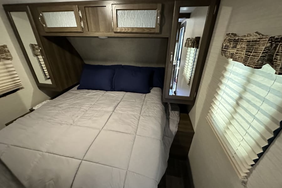 RV rental bedroom: cozy space with bed, blue pillows, overhead cabinets, mirrored doors, and pleated window blinds.