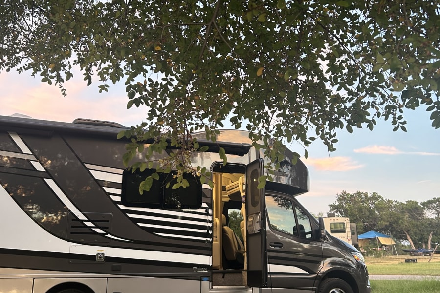 An RV rental with its door open is parked on gravel under a tree, with sky and distant tents visible. Ideal for camping.