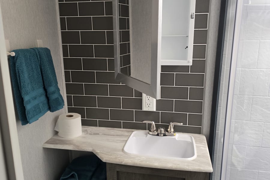RV rental bathroom featuring a vanity sink, open medicine cabinet, dark tiled backsplash, teal towels, and a shower.