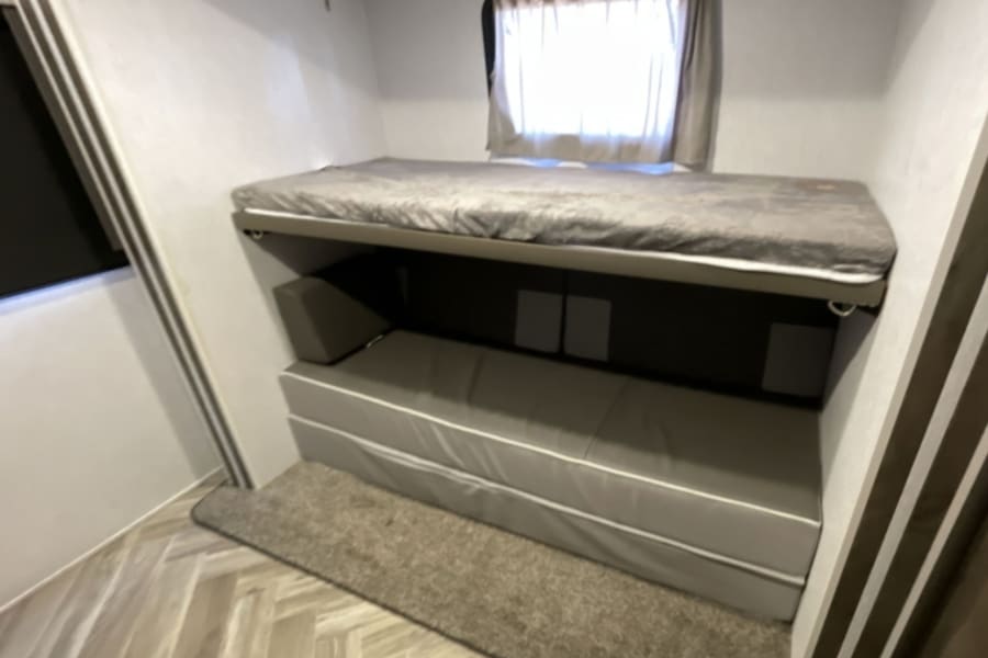 Inside an RV rental: a cozy bunkhouse with gray bunk beds, bright window, and light wood herringbone floor.