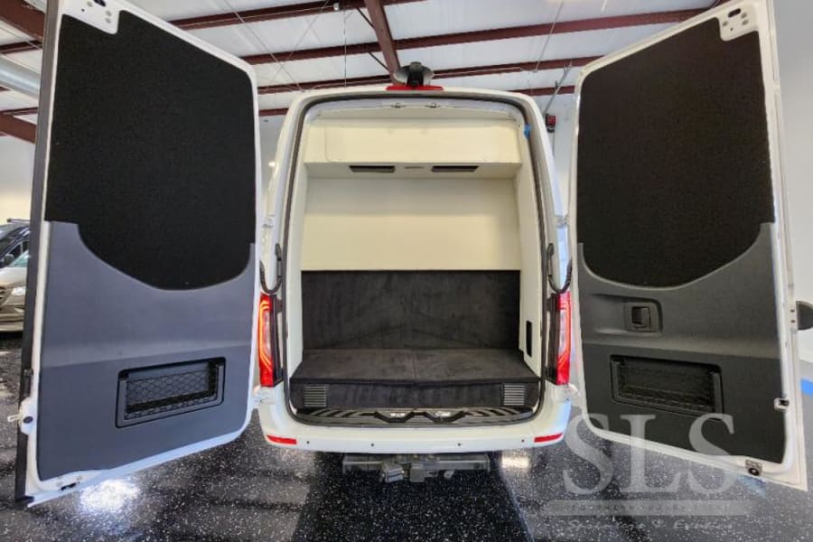 Rear view of an open white camper van for RV rental, showing its custom interior with a bed/bench and door storage.
