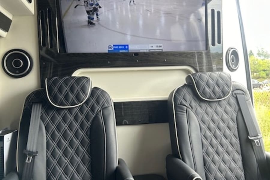 Inside a luxury **RV rental**: two plush captain's chairs, central console, and a large TV showing a hockey game.