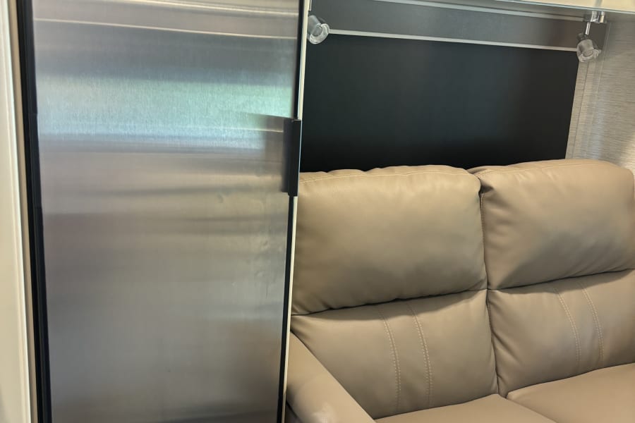 Modern stainless steel refrigerator and light-colored leather seating in a clean rv rental interior, with overhead cabinets and lighting.