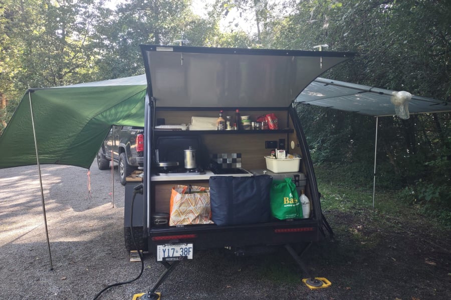 This RV rental trailer features a convenient outdoor kitchen under extended awnings at a quiet campsite, perfect for glamping.