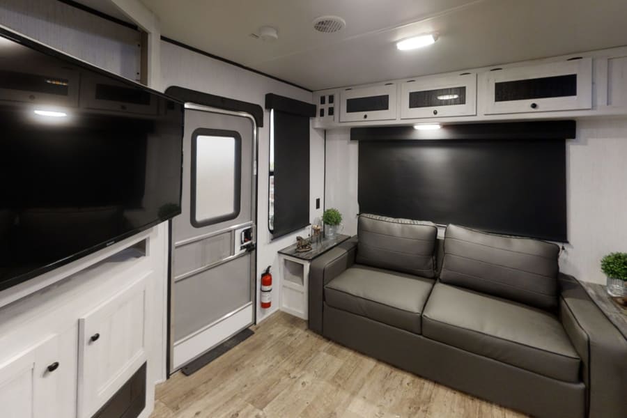 Inside a modern RV rental: Living space with a TV, grey sofa, overhead cabinets, and an entry door. Brightly lit.