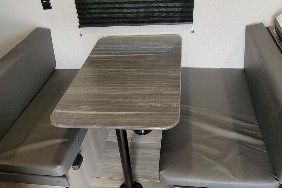 **RV rental** dinette: gray benches, wood-grain table, window with blind, light walls & floor, colorful rug.