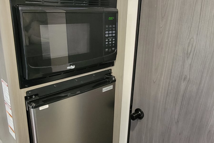 Inside an RV rental, featuring a streamlined kitchen with a black microwave, stainless steel fridge, and light wood cabinets.