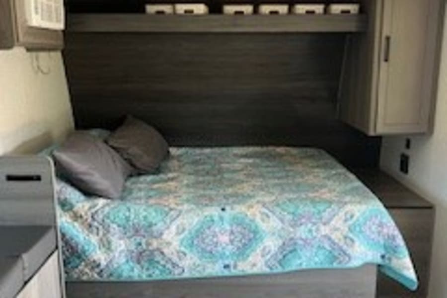 RV rental bedroom: a comfy bed with blue quilt, overhead shelving, side cabinets, and an AC unit on the left.