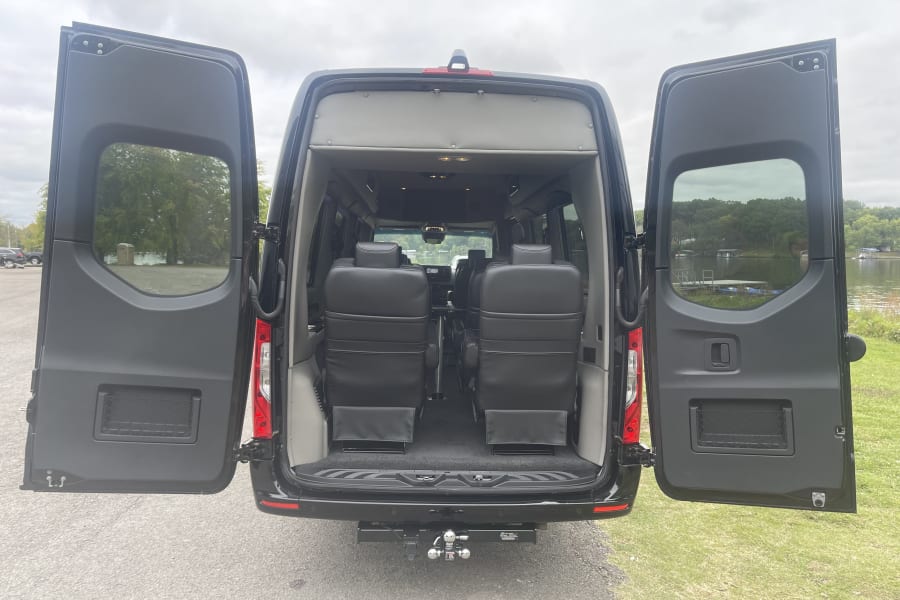 This black luxury RV rental displays its open back doors, revealing comfortable interior seating, a tow hitch, and a scenic lake view.