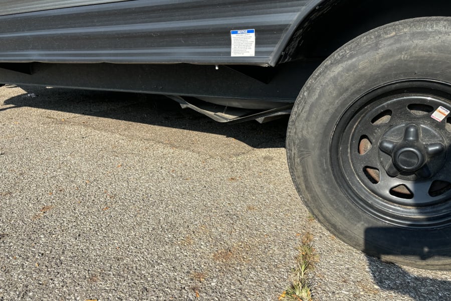 A close-up of an RV rental trailer's side and wheel on asphalt, featuring a safety notice about checking wheel lug nuts.