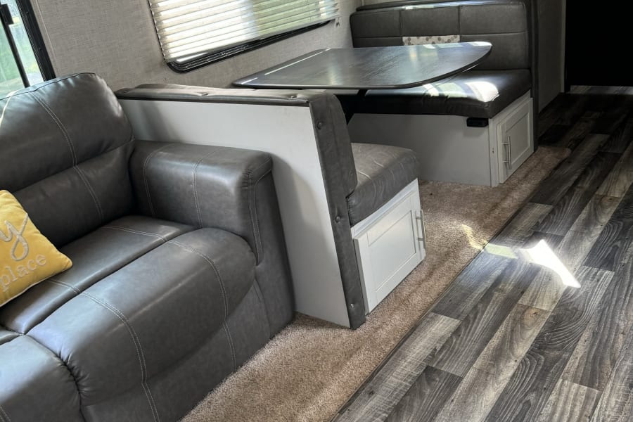 Spacious rv rental interior featuring a gray sofa, dining booth with table, wood-look flooring, and a beige carpeted area.