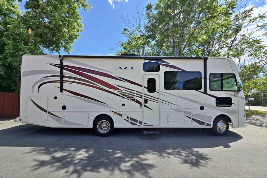 Side view of a large cream "ACE" Class A motorhome with burgundy/gray graphics, parked on asphalt, perfect for an RV rental.