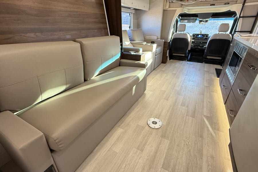 This RV rental's modern motorhome interior features a cozy sofa, lounge chairs, and a kitchen, leading to the cockpit.