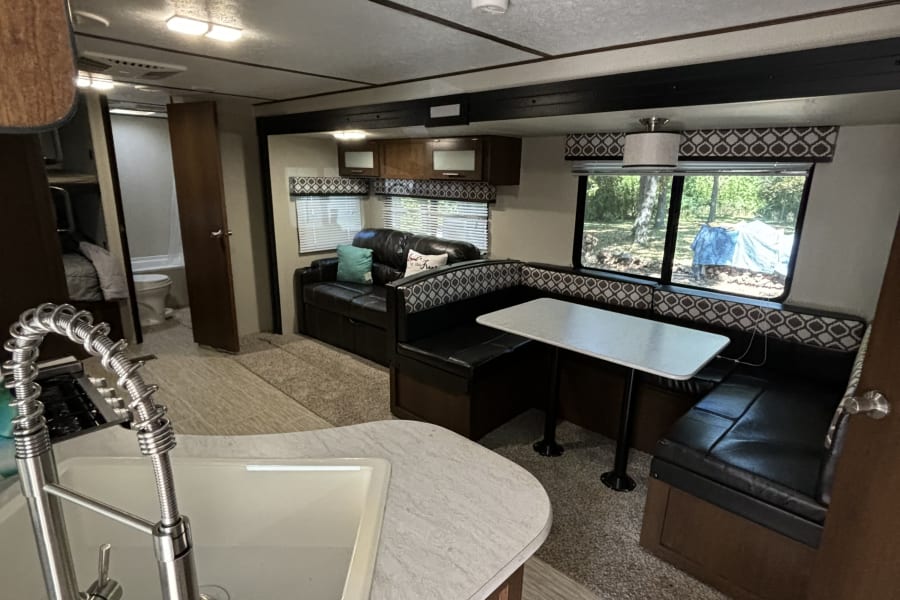 Spacious RV rental interior featuring a dining booth, sofa, and kitchen sink with a view of nature outside.
