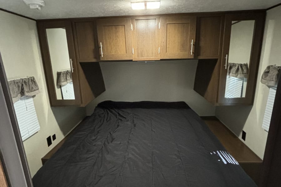 Cozy RV rental bedroom featuring a dark bed, wooden overhead cabinets, and windows with blinds.