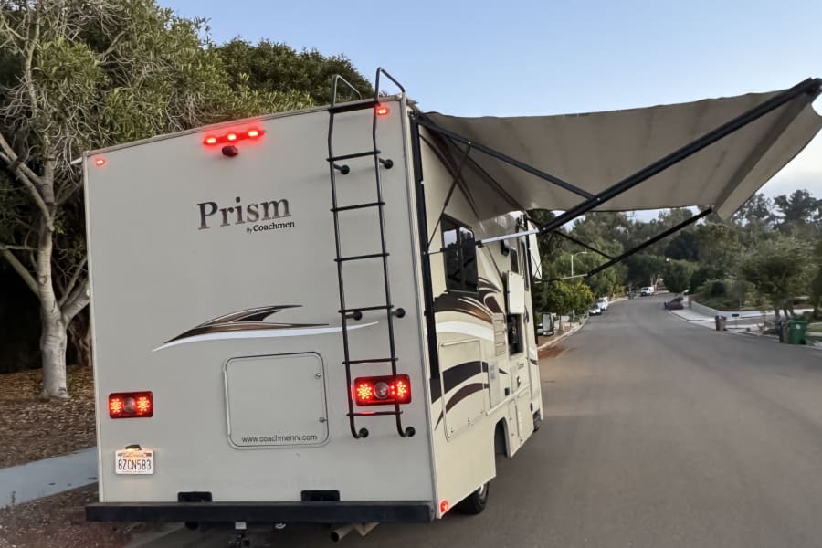 RV rental: A beige Coachmen Prism motorhome with its awning extended and taillights on, parked on a street.