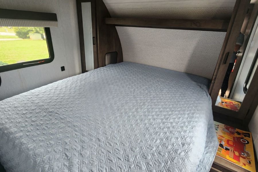 Cozy RV rental bedroom with a light blue quilted bed, window offering green views, and practical built-in storage for comfortable travel.