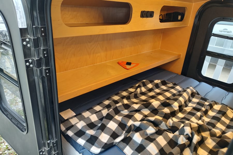 Explore this RV rental's comfy interior: a bed with checkered blanket, built-in wooden storage, and a large window.