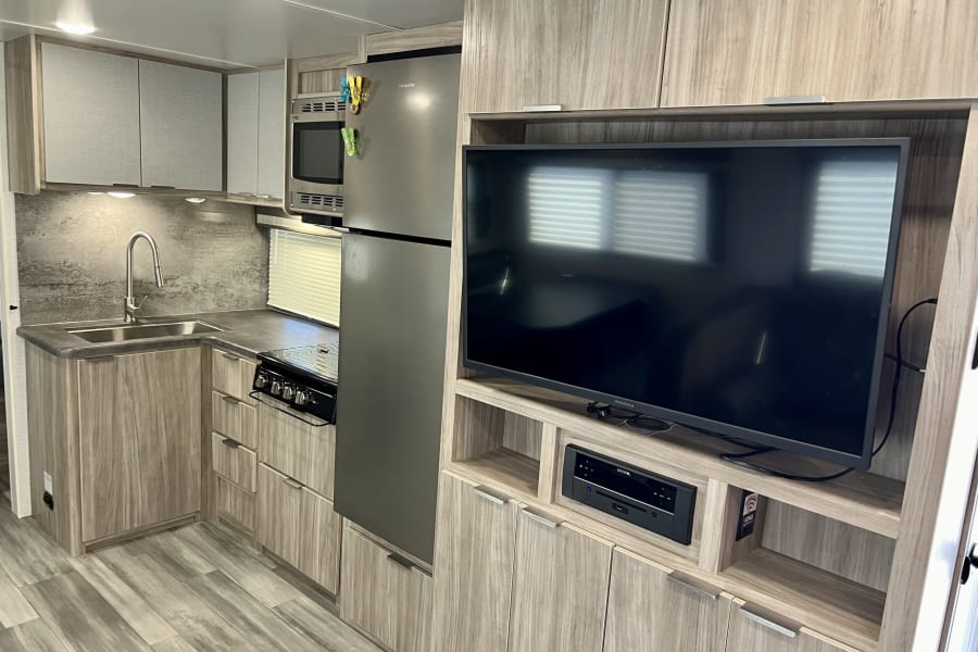 Modern rv rental interior: kitchen with sink, stove, microwave, stainless fridge, and entertainment center with TV.