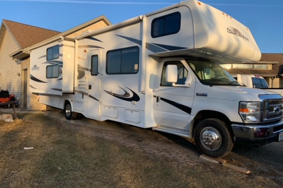 A white Sunseeker Class C RV with extended slide-out, parked in a driveway next to a house. Great for an RV rental adventure.