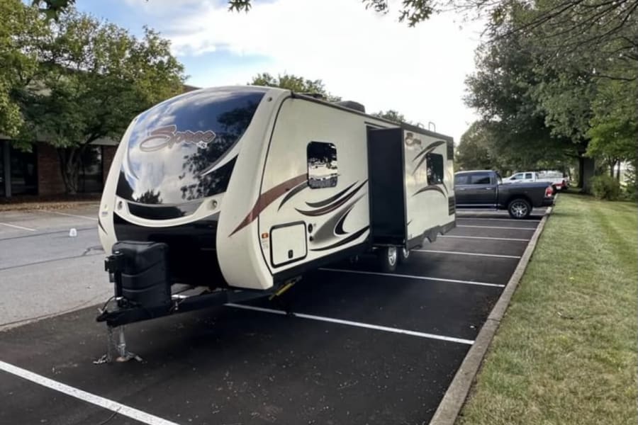 Spacious cream travel trailer (RV rental) with slide-out, parked in a lot under trees.
